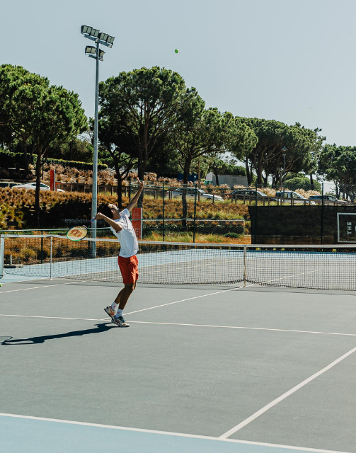 Um homem está num campo de ténis num campus, a preparar-se para servir a bola durante um jogo.