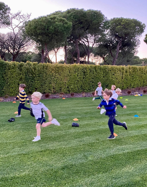 Crianças a jogar futebol num campo relvado no campus, a desfrutar de um dia de sol com atividades ao ar livre.